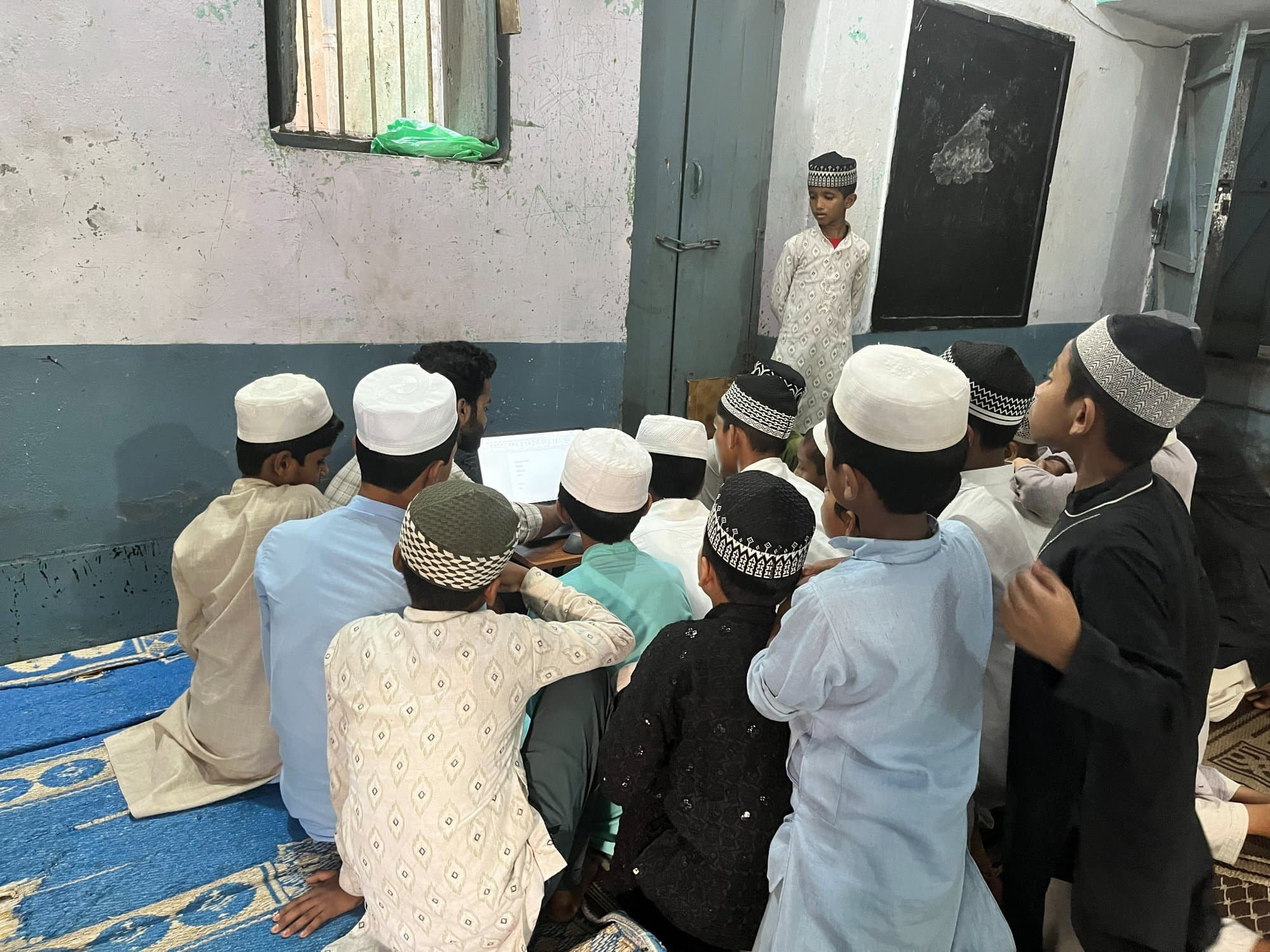 मदरसे की कक्षा में स्वयंसेवक के नेतृत्व में सत्र, साझा लैपटॉप के चारों ओर जुटे छात्र।